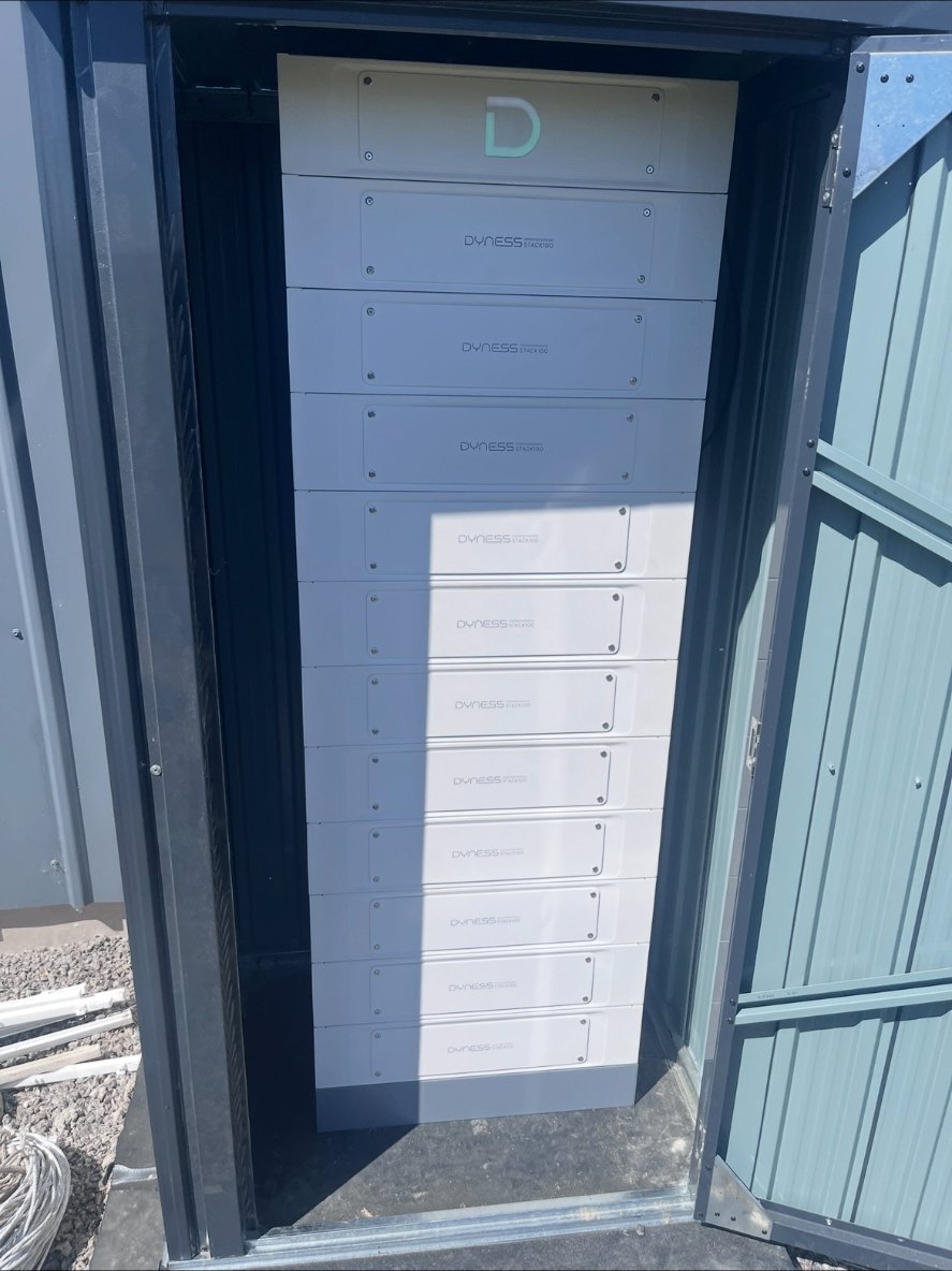 Högt, vitt batteripack i tornformat installerat inuti ett mörkt plåtskåp utomhus vid en villa i Norrköping, med kabelkanaler på grusmarken intill